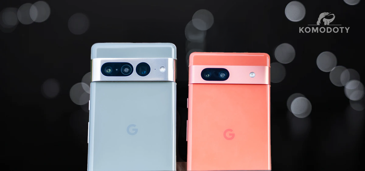 Side-by-side view of the Google Pixel 9a in gray and Pixel 7a in coral, showcasing rear camera designs and hardware differences against a blurred dark background with Komodoty branding.