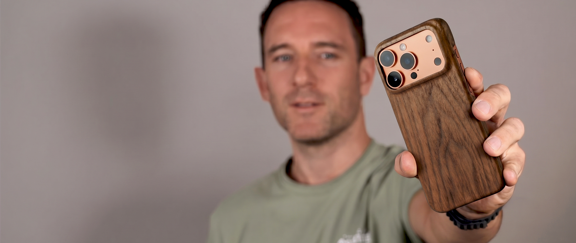 caucasian Man in a light green tshirt holding a smartphone with a slim wooden case for his iphone 17 pro max dessert orange against a plain beige background in a studio