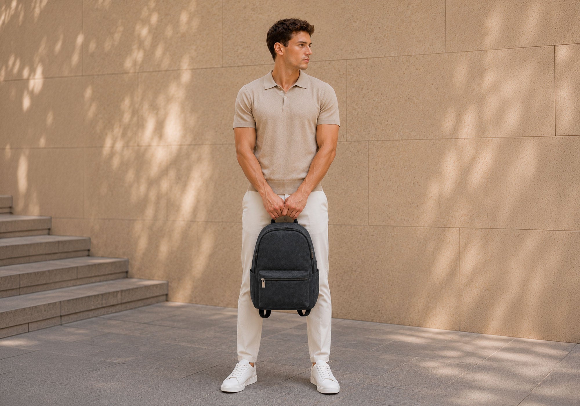 Male model in a beige collared shirt with white pants holding a black alcantara backpack against a beige wall outdoors