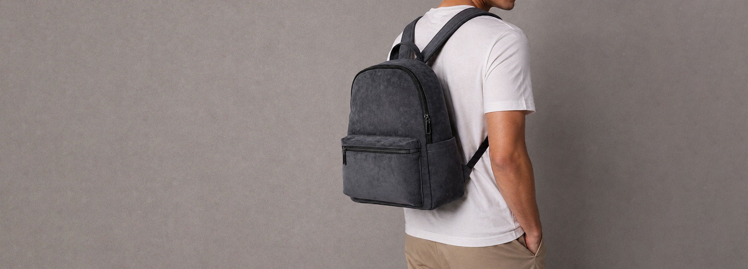 Male model in a beige collared shirt with white pants holding a black alcantara backpack against a grey studio background