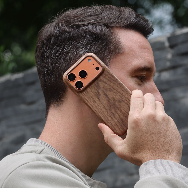 Komodoty Slim Wood iPhone Case on an iPhone 17 Pro Orange held on a man's ear with a blurred background in the park