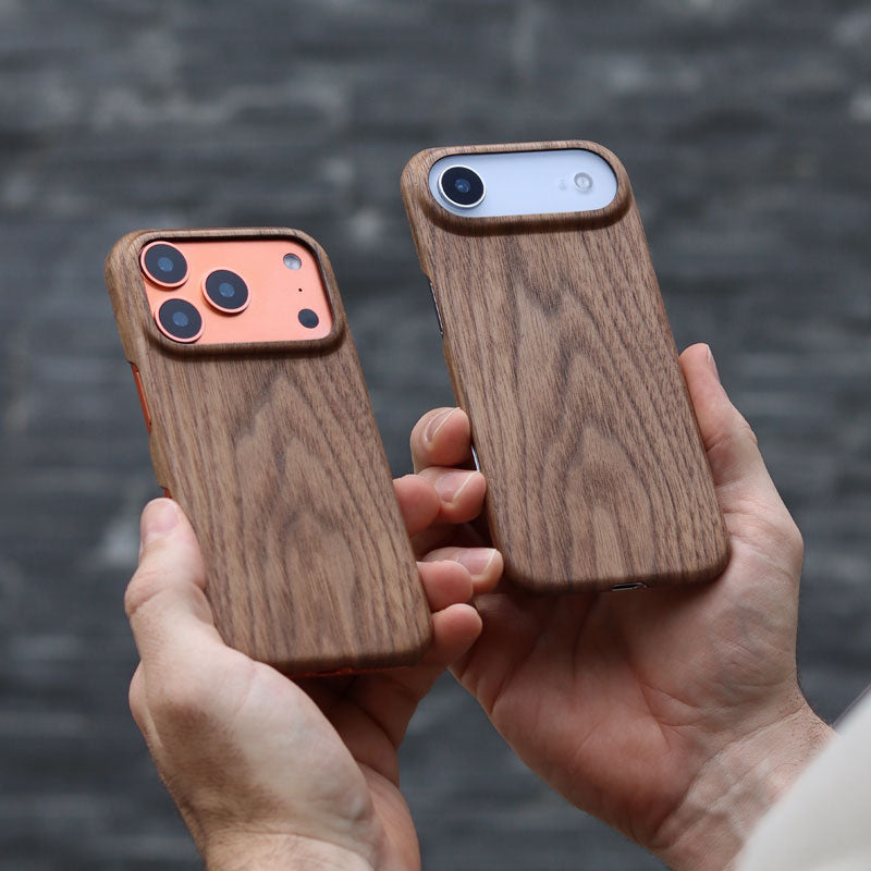 Two wooden phone cases for iPhone 17 Pro and iPhone Air held by hands against a blurred grey brick wall background