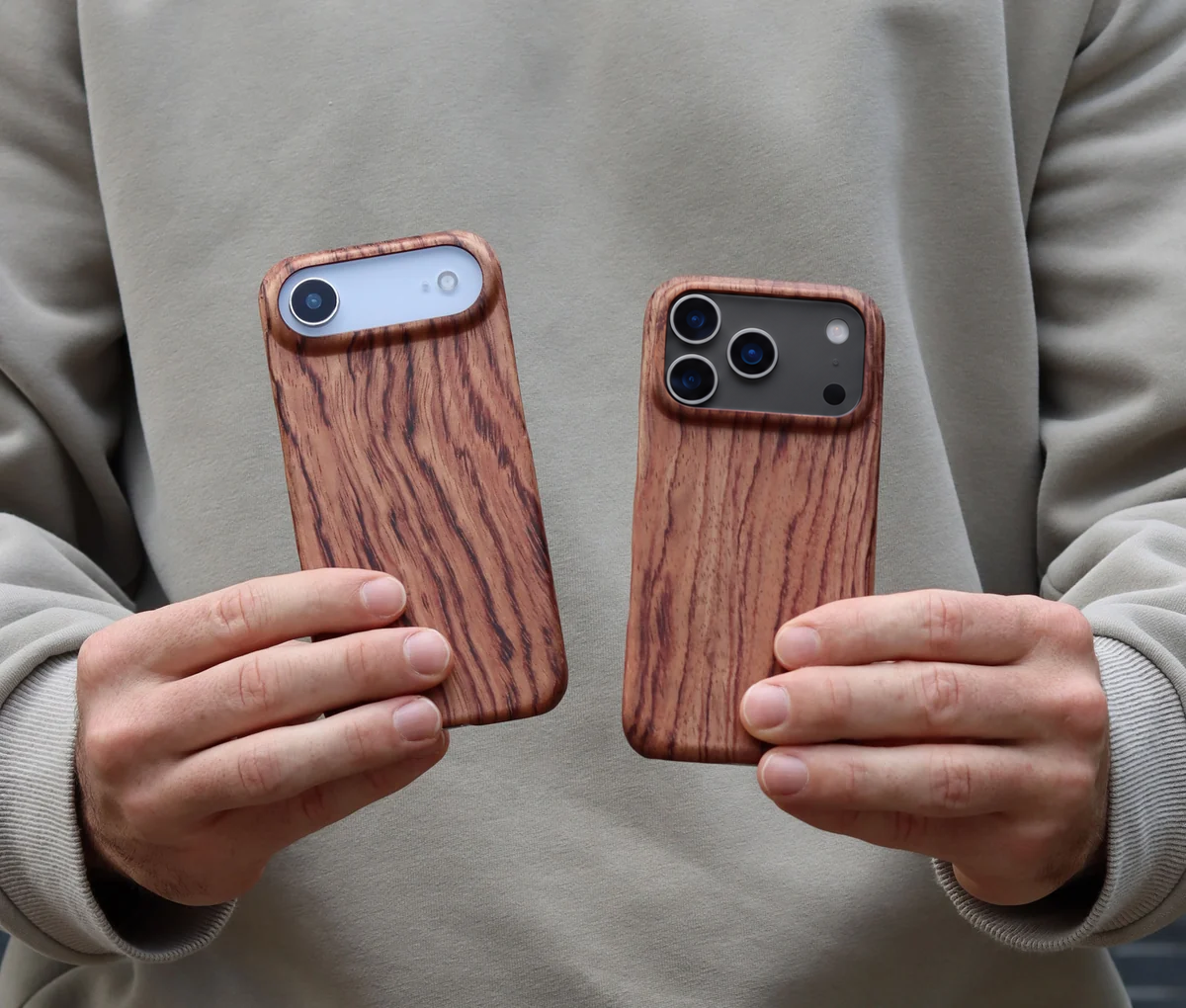 Komodo two iPhones held by a model left is iPhone Air in White and right is the new iPhone 18 Pro Max in Black both with a rosewood wooden phone case, background is a man's beige northface sweater.