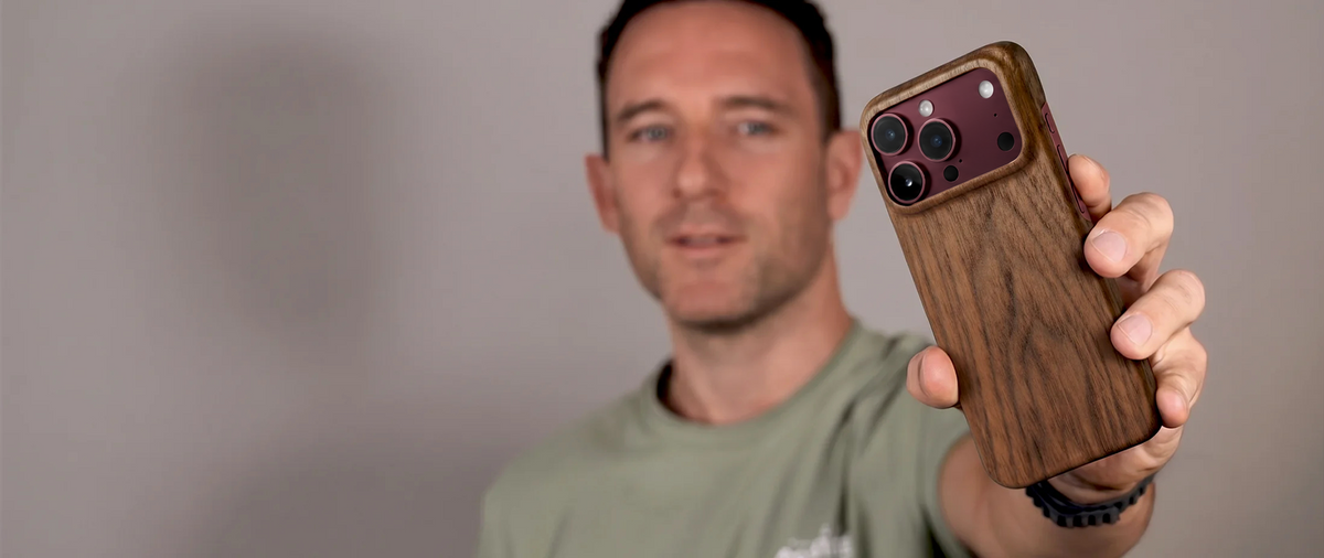 caucasian Man in a light green tshirt holding a smartphone with a slim wooden case for his burgundy iphone 18 pro max dessert orange against a plain beige background in a studio