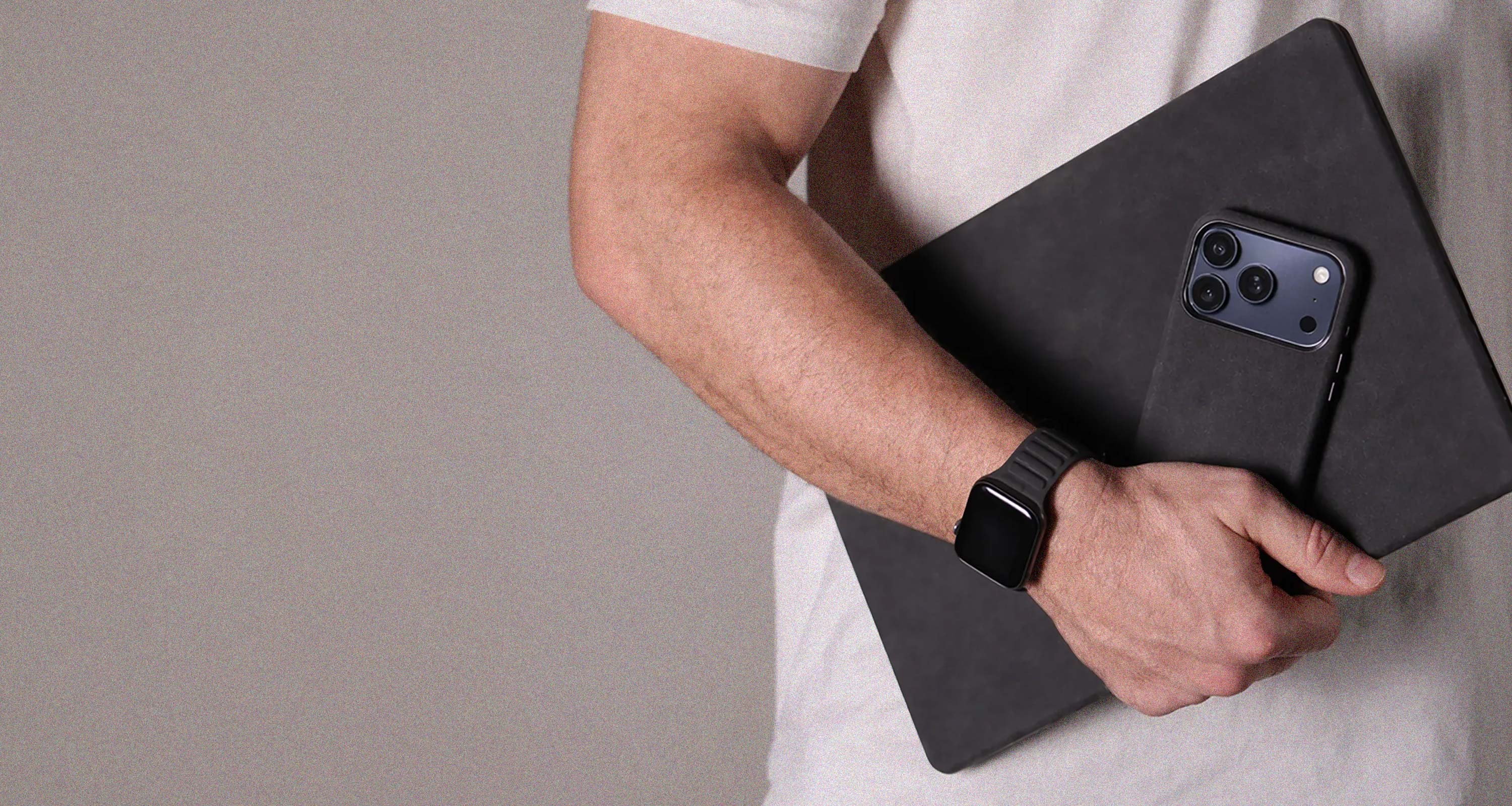 Person holding a black alcantara laptop case for macbook pro with a black alcantara case on a iphone 17 pro max blue and wearing a black alcantara apple watch band on a plain background