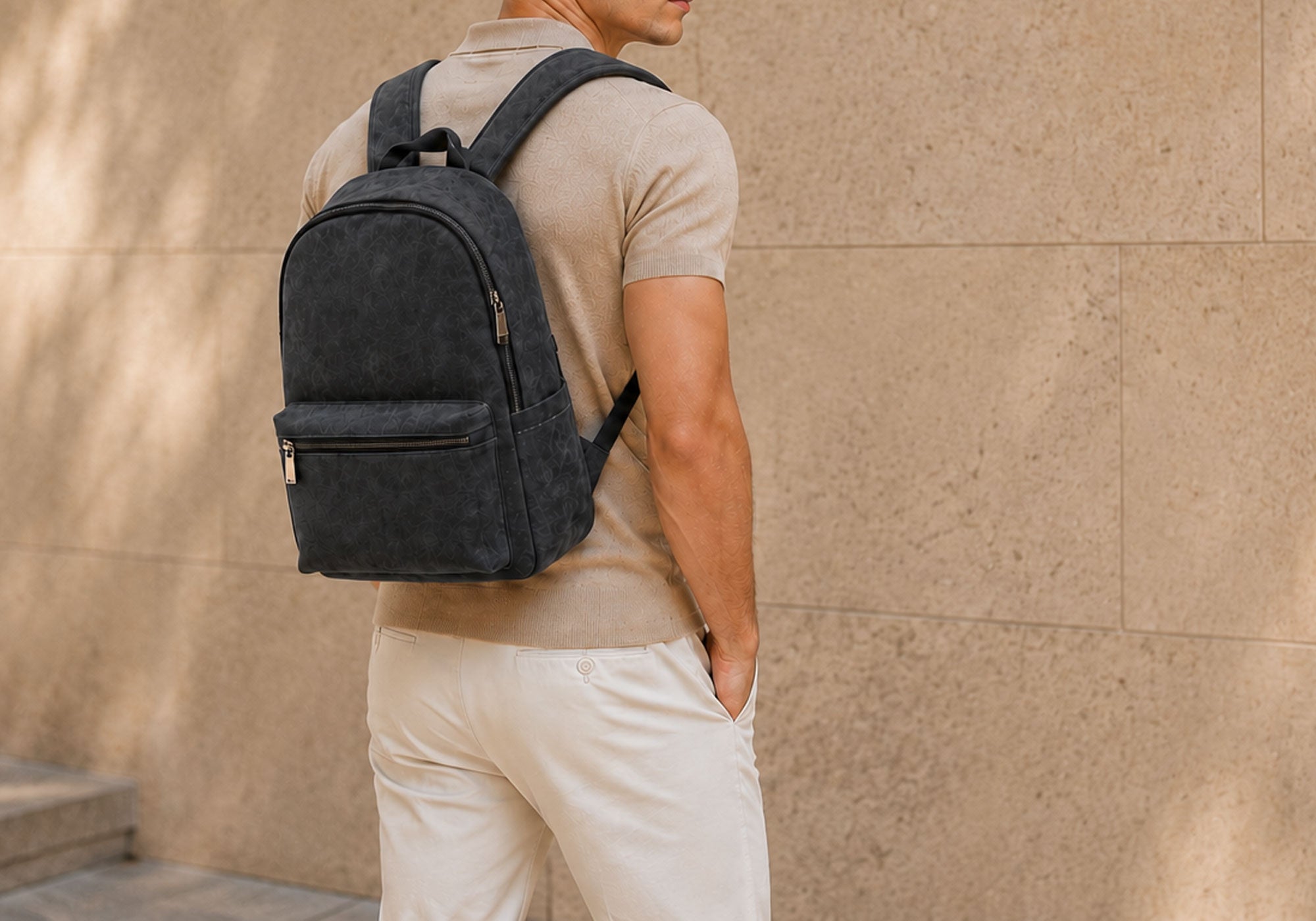Male model in a beige collared shirt with white pants wearing a black alcantara backpack against a beige wall outdoors