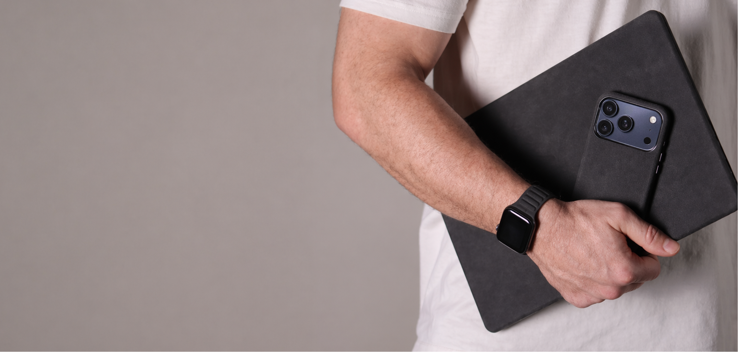 Man holding a black alcantara suede like cover on a macbook pro with a matching black alcantara iPhone 17 Pro Max on a plain background
