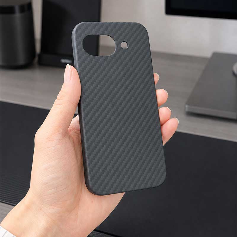 A hand holding a black carbon fiber textured phone case for Google Pixel 10A with camera cutouts, set against a blurred desk background.