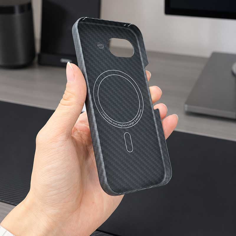 A hand holding a black carbon fiber pattern phone case for Pixel 10A with a circular MagSafe ring design, shown against a blurred desk background.