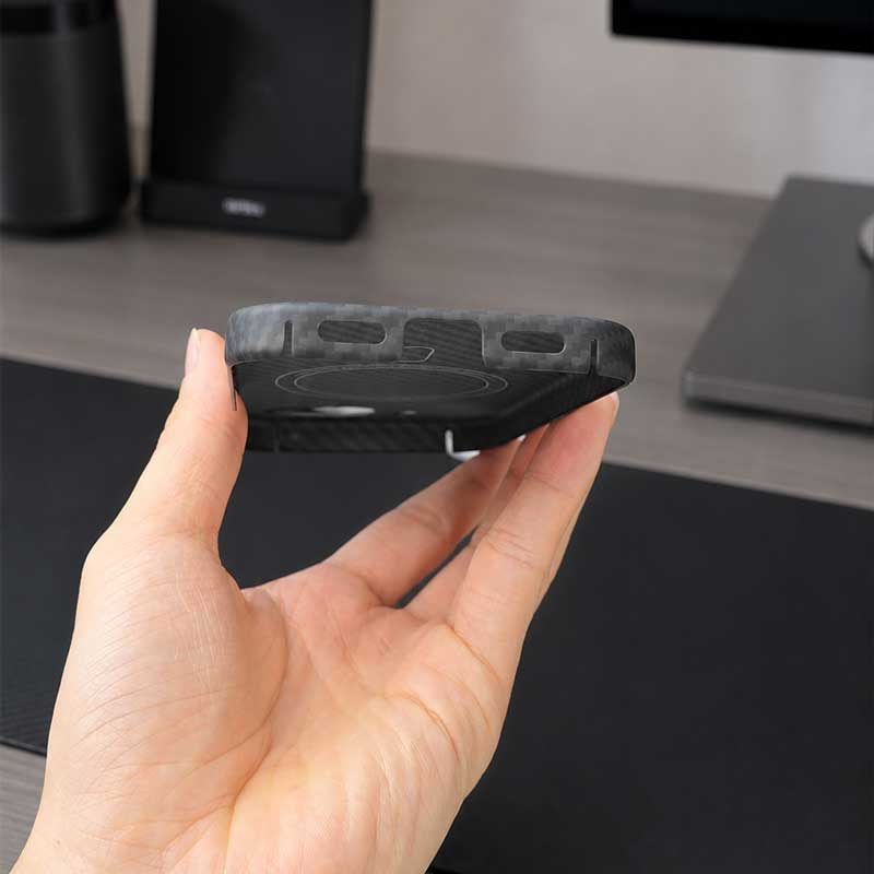 A hand holding a black rugged phone case for Pixel 10A with a carbon fiber pattern, shown in a close-up shot against a blurred desk background.