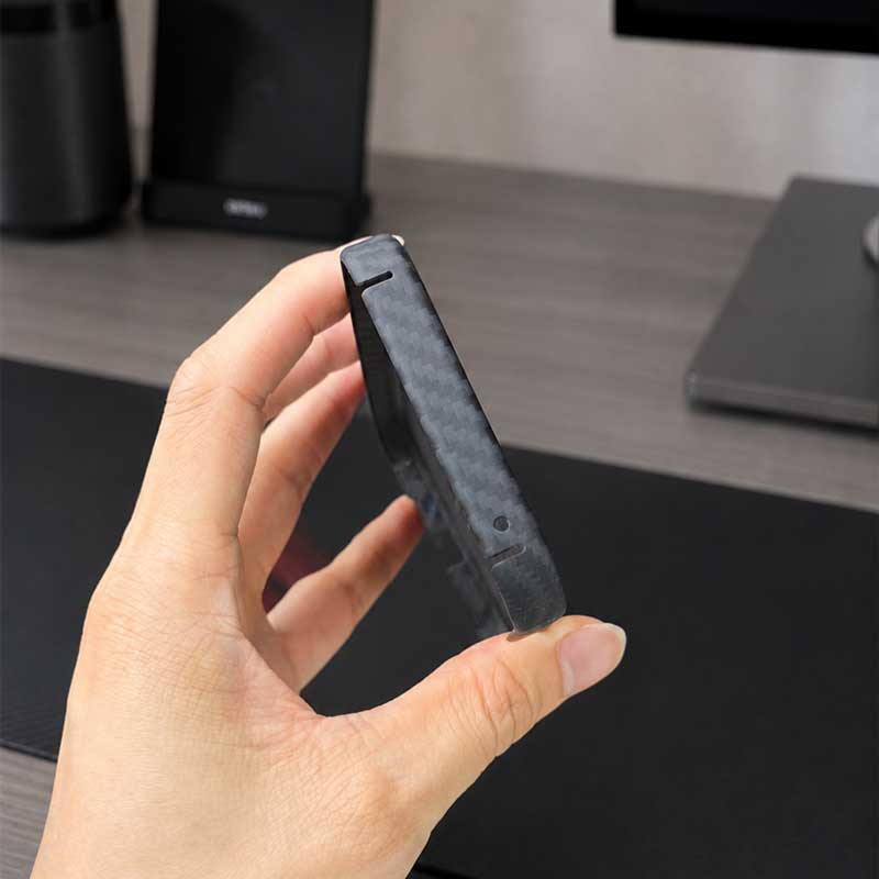 A hand holding a Pixel 10A carbon fiber phone case, shown in a close-up against a blurred desk background.