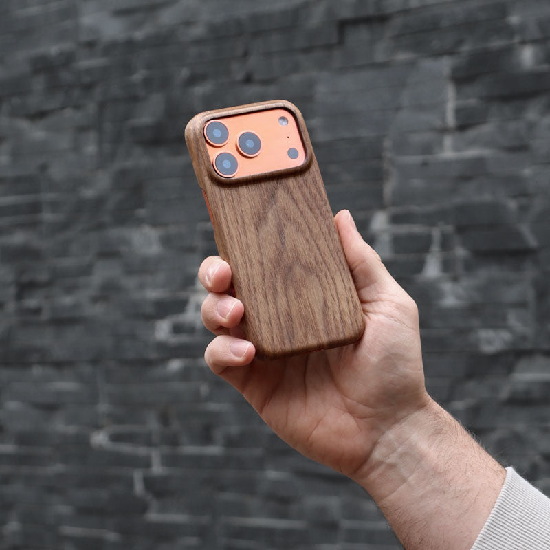 Komodoty Slim Wood iPhone Case on an iPhone 17 Pro Orange held by man's hand upright with slight angle against a grey stone wall.