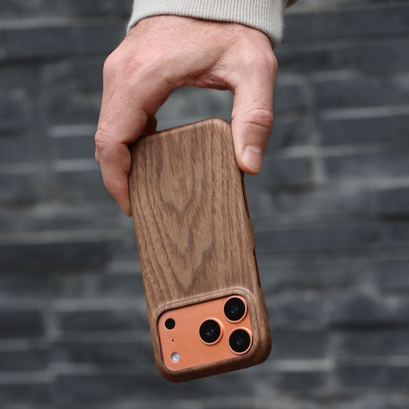 Komodoty Slim Wood iPhone Case on an iPhone 17 Pro Orange held by man's hand upside down against a grey brick wall.
