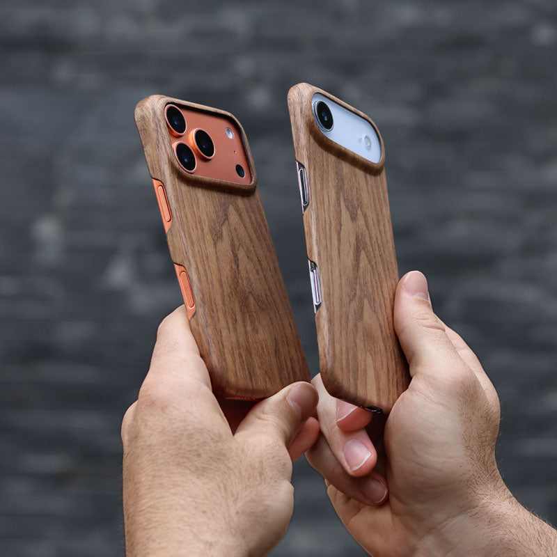 Two hands holding two iPhone 17 Pro and iPhone Air in Komodoty's Slim Wood iPhone case against a dark grey brick wall background