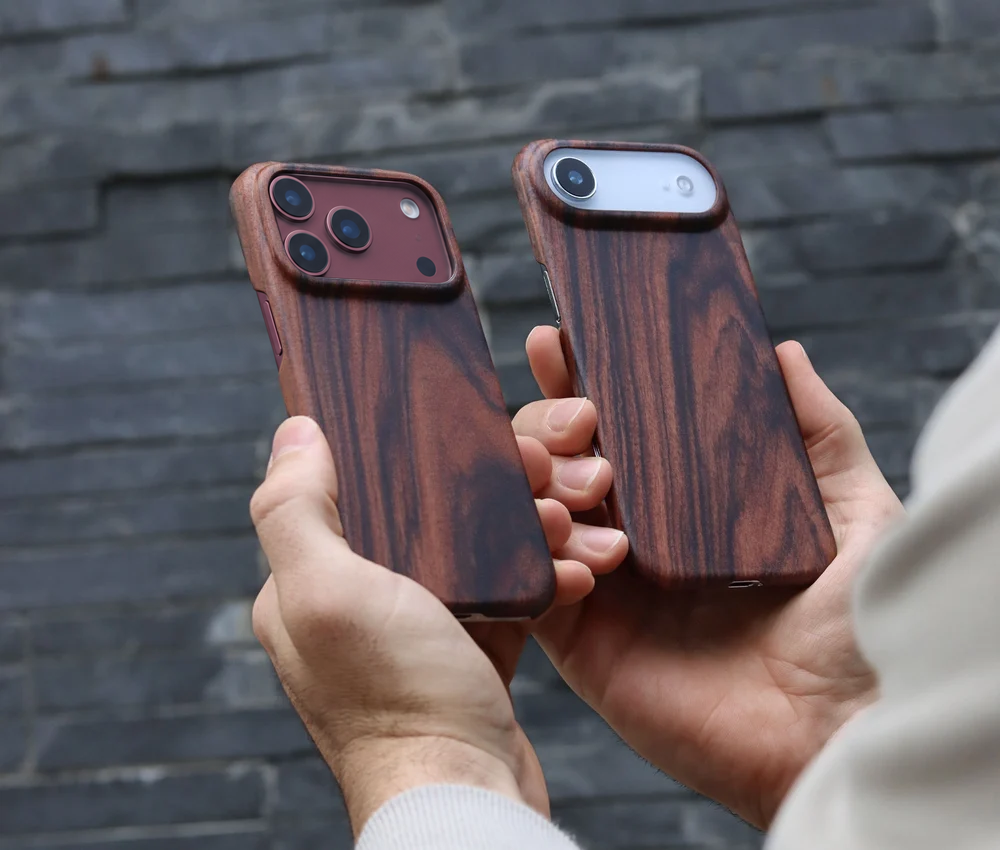Two hands holding two wooden iphone cases with a rich dark brown grain in Mahogany wood, left is the iphone 18 pro in burgundy and the right is the iphone air in white shown against a dark gray brick wall background.